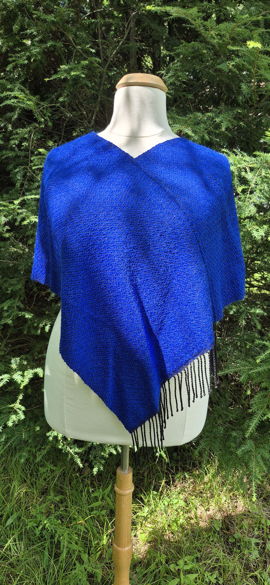 Blue alpaca and Tencel poncho with fringe on a dress form, outdoors with plants behind