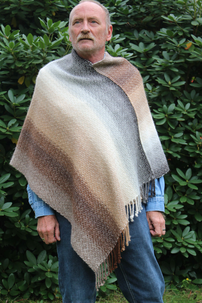 Man dressed in denim, wearing a natural alpaca Mexican poncho in shades of brown and gray, a large green bush behind him